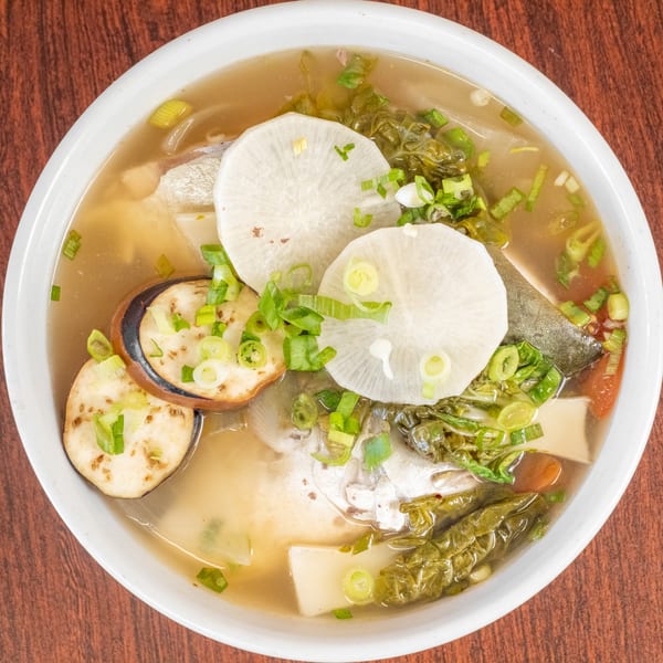 A white bowl of fish soup with eggplant, radish, and green onions on a wooden table.