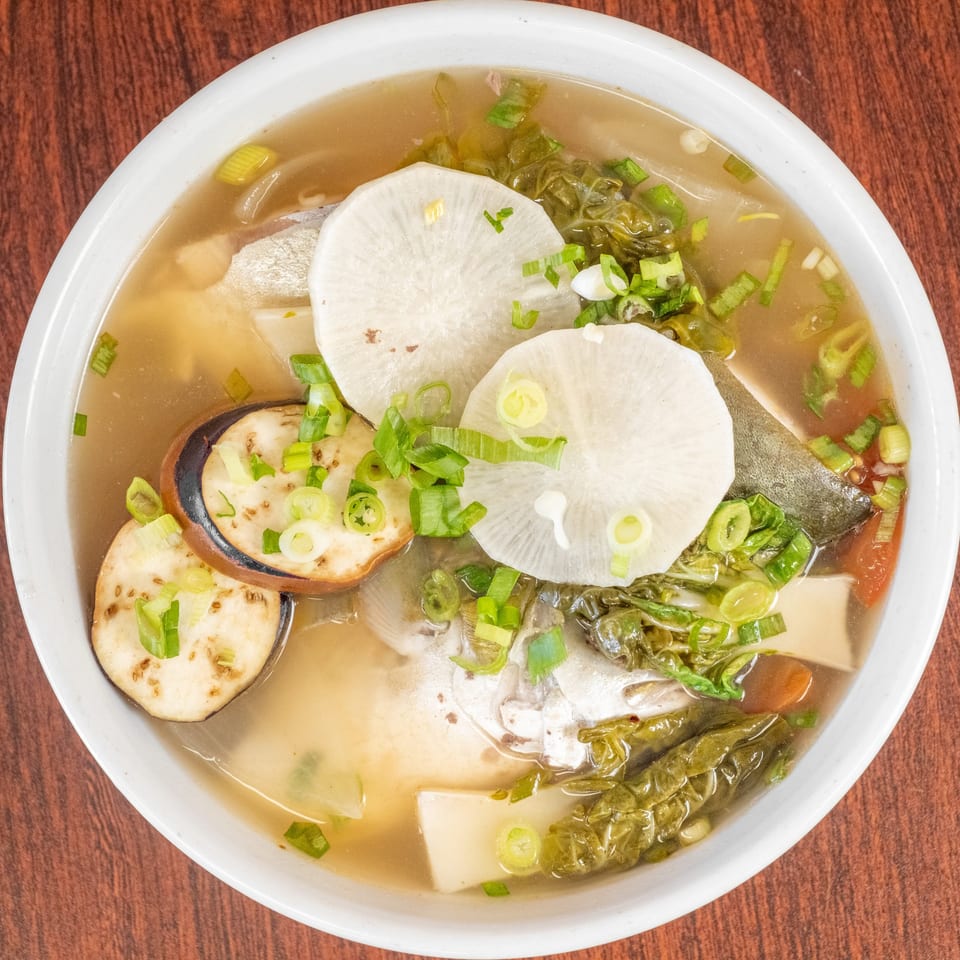 A white bowl of fish soup with eggplant, radish, and green onions on a wooden table.