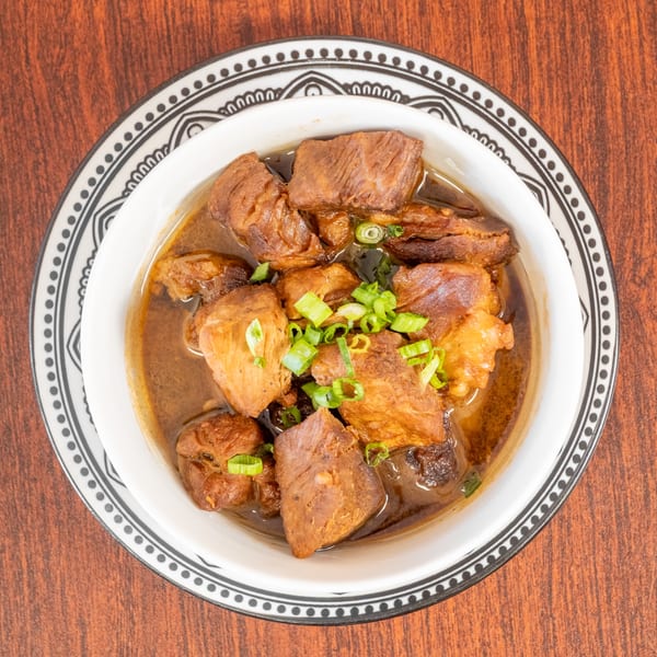 Braised pork belly with dark sauce and chopped green onions in a white bowl with a patterned rim.