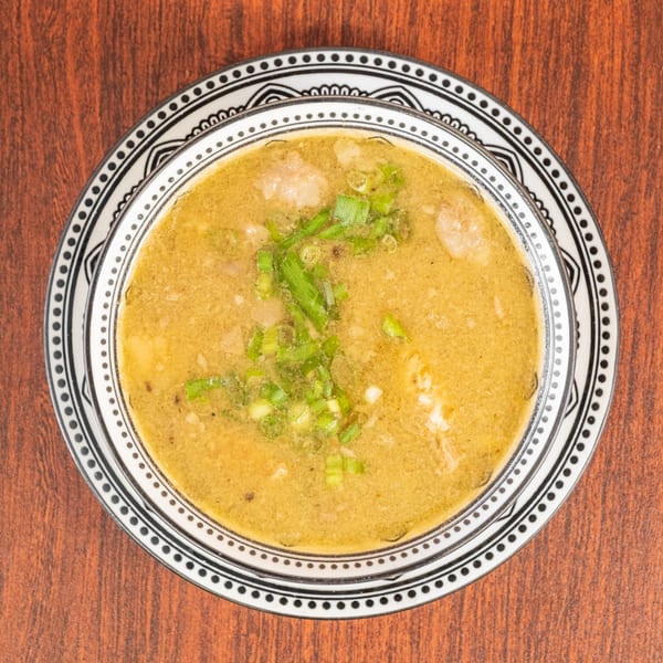 Overhead view of a bowl of green curry with chicken and chopped scallions on a wooden table.