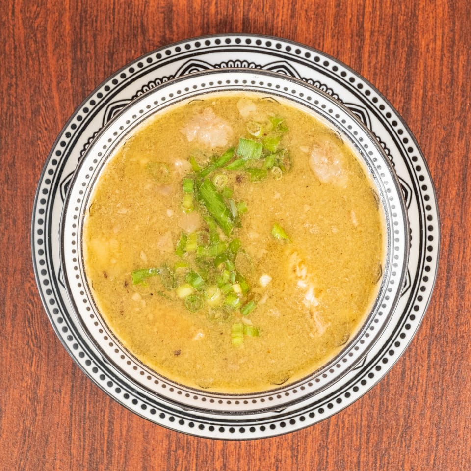 Overhead view of a bowl of green curry with chicken and chopped scallions on a wooden table.