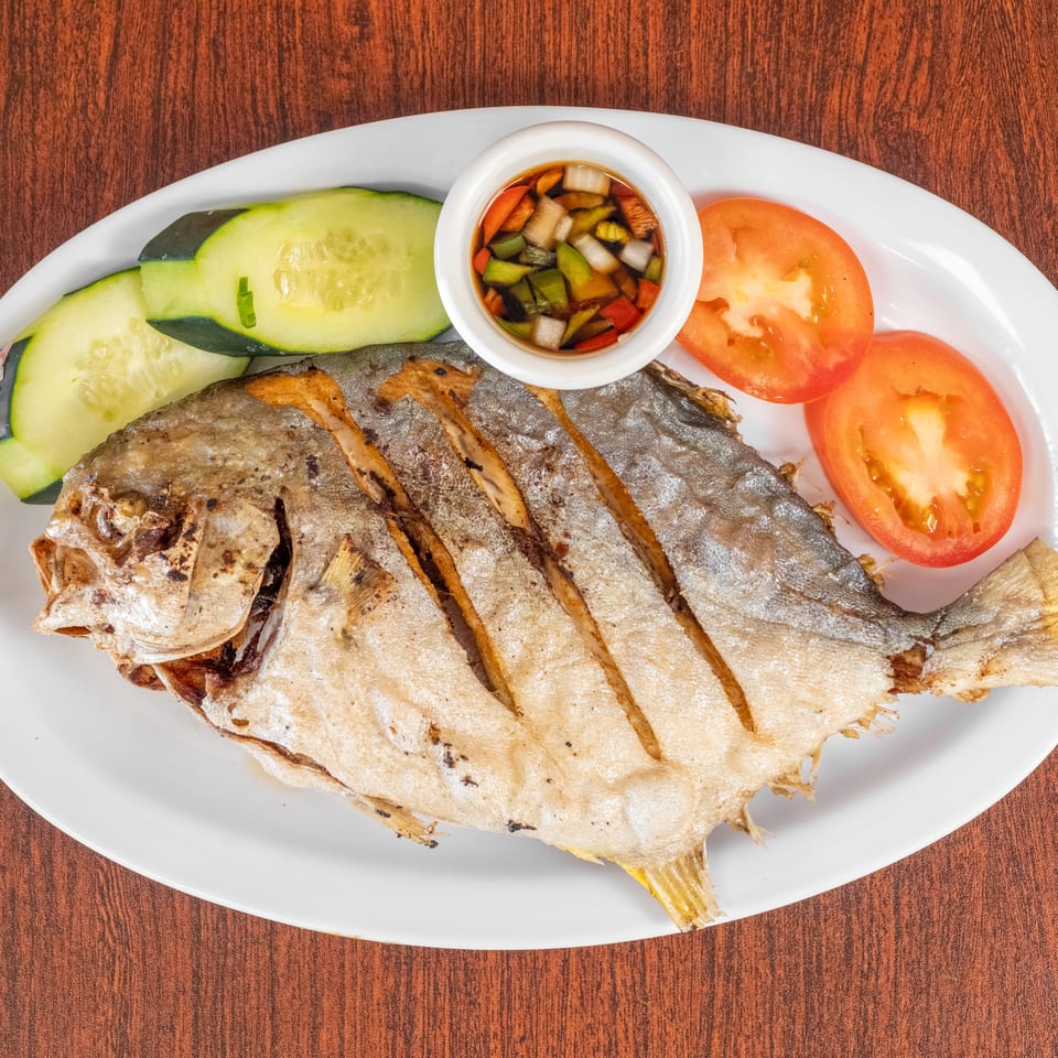 Grilled pompano fish with sliced cucumbers, tomatoes, and a dipping sauce on a white plate.