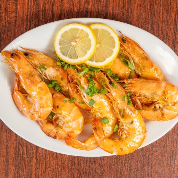 A white oval plate filled with large, cooked shrimp garnished with lemon slices and chopped green onions.