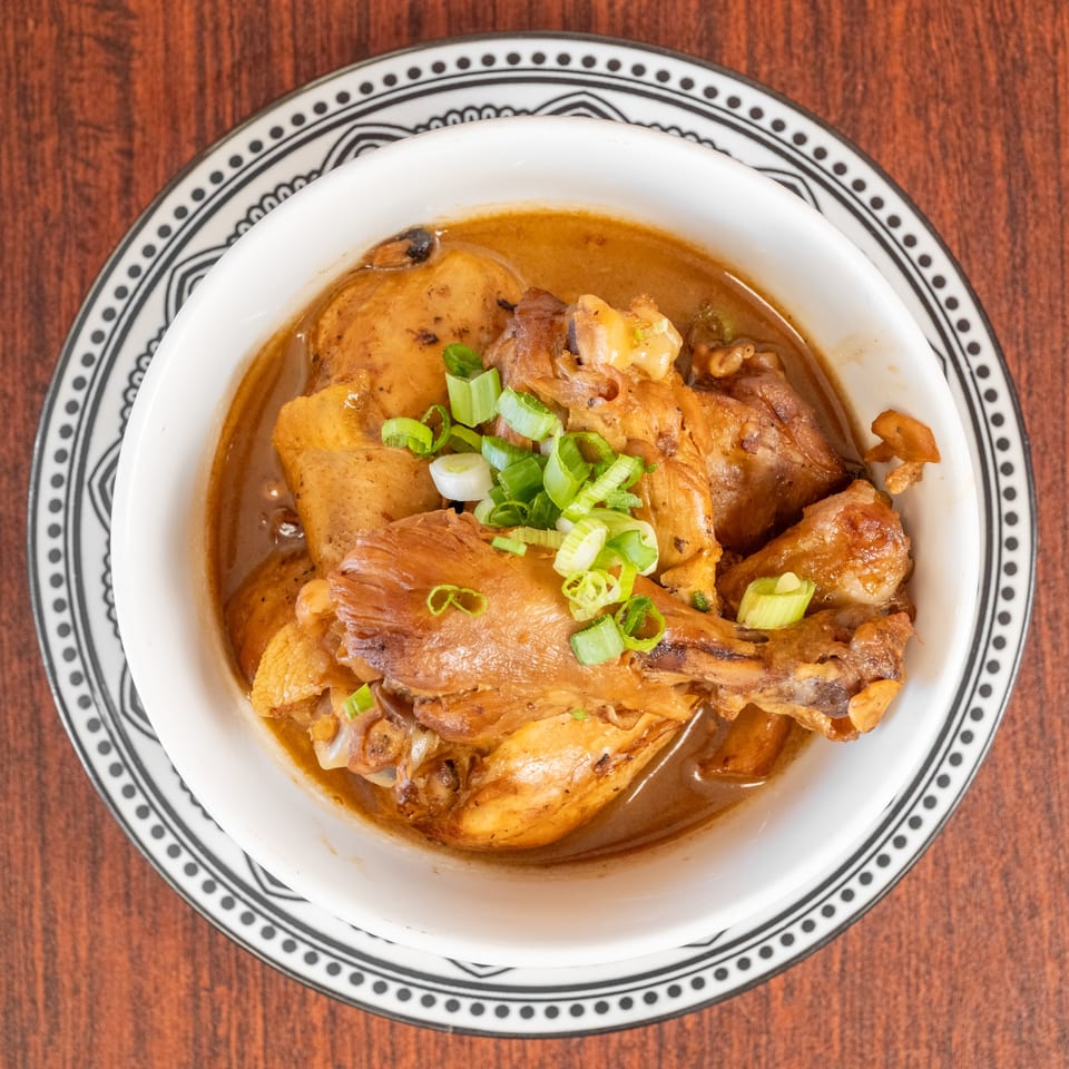 Chicken adobo with potatoes and green onions in a white bowl with a patterned rim.